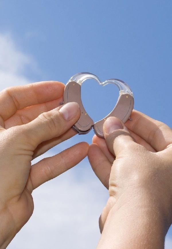 Zwei Hände formen mit Hörgeräten ein Herz vor blauem Himmel zur Hörgeräteversorgung.